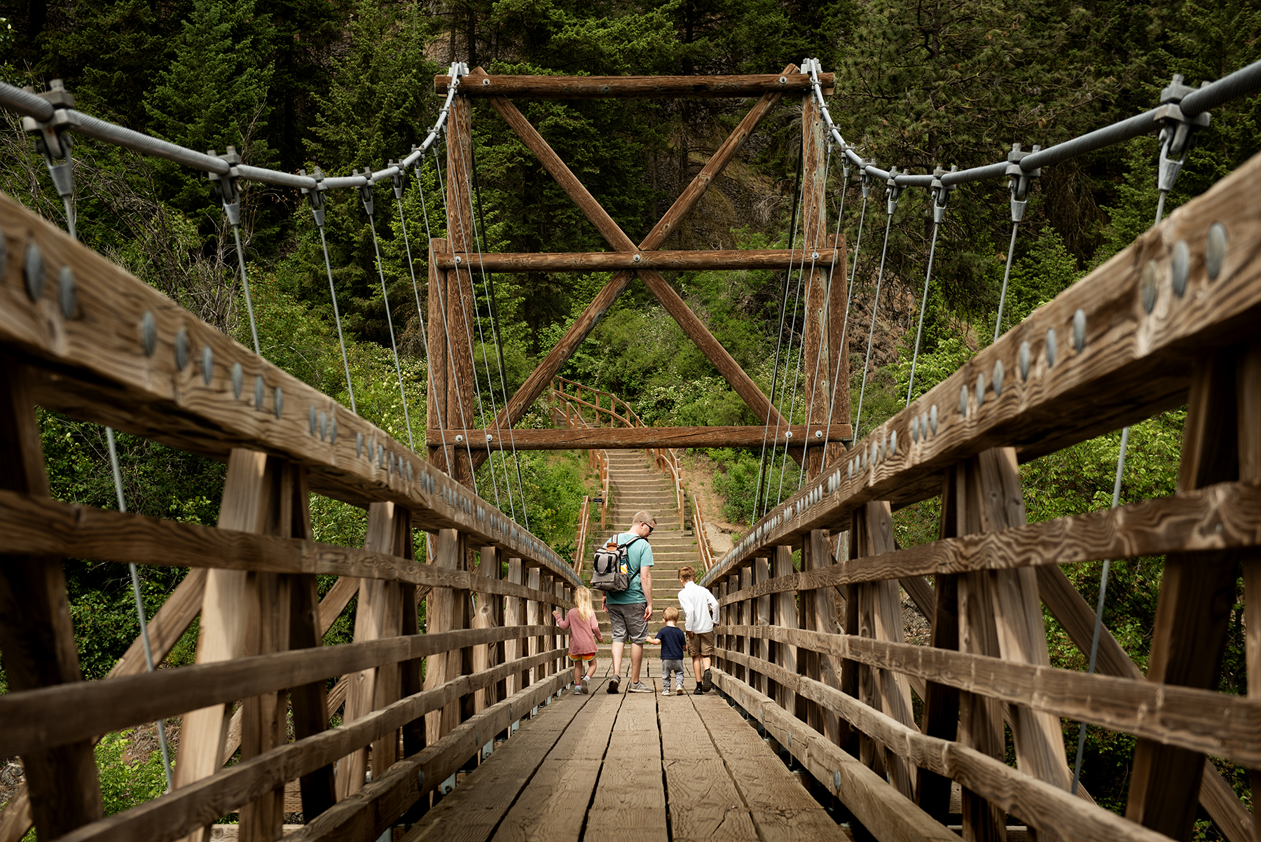Family of one adult and three small children crosses a suspension bridge, headed into the forest