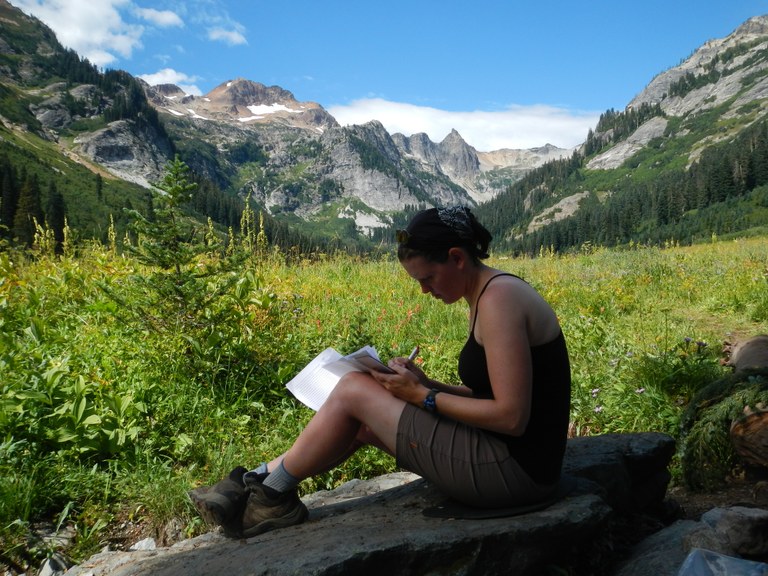 A hiker sits on a rock and writes in a journal. 
