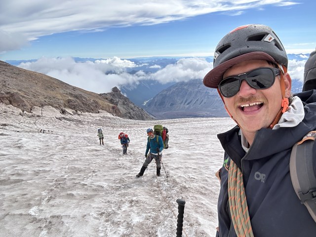 A guide wearing a helmet and sunglasses leads a group of three climbs up Mount Rainier.