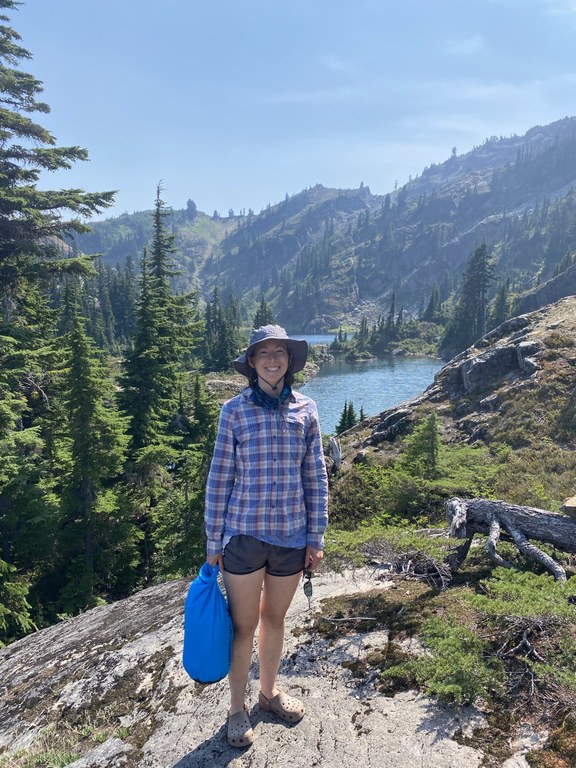 Kyli stands on a rocky outcropping above a lake, smiling a the camera with a large stuff sack in one hand.