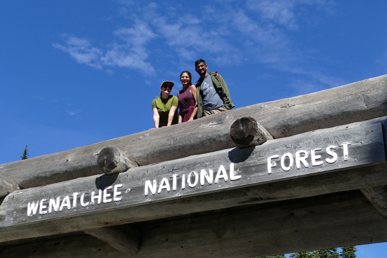 Three hikers in their late 20s smile at the camera on top of a bridge with a "Wenatchee National Forest" sign with a backdrop of bright blue sky. 