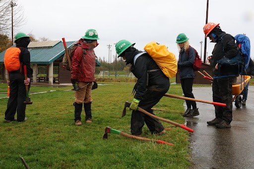 Five volunteers in hard hats wait in line to grab tools. One person in green hat with bright yellow rain cover on bag bends down to grab tool. Park shelter in background with green grass on a grey, rainy day.
