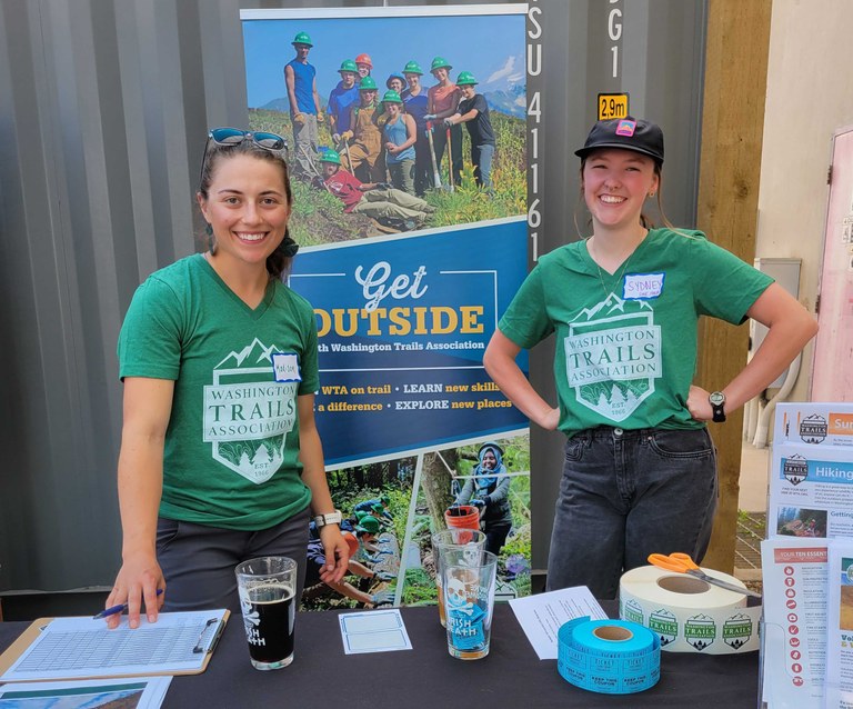 Two WTA employees in WTA shirts stand a table in front of a sign saying "get outside."