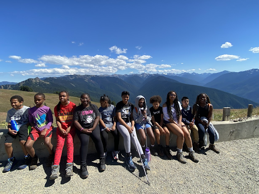 Group of 11 young hikers/campers smile in front of blue skies and mountains in the background while sitting against a short concrete wall. 