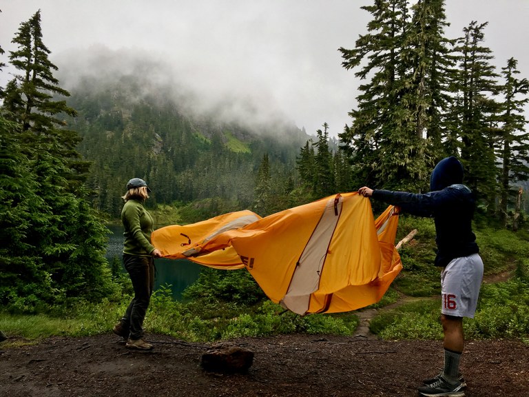 Two hikers spread out their tent in camp with a foggy lake in the background.