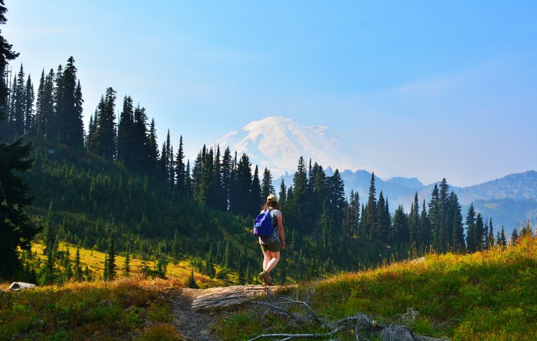 A hiker strides across the trail with Mount Rainier in the background.