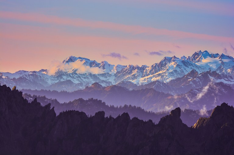 Layers of mountains, with snowy peaks in the background and a purple sunset. 