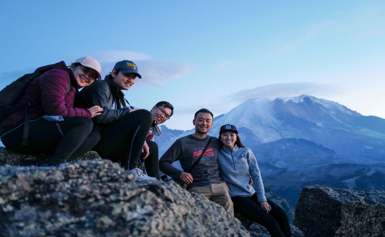 A group of hikers arranged on a rock smile at the camera, with Mount Rainier behind.