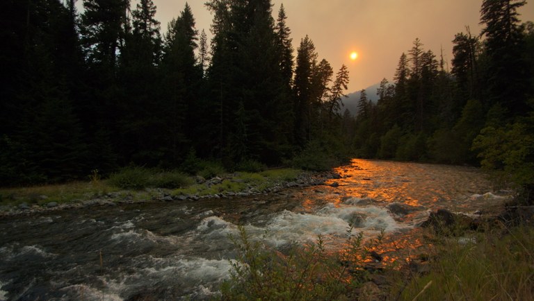 An orange sun shines in a smoky sky over a river.
