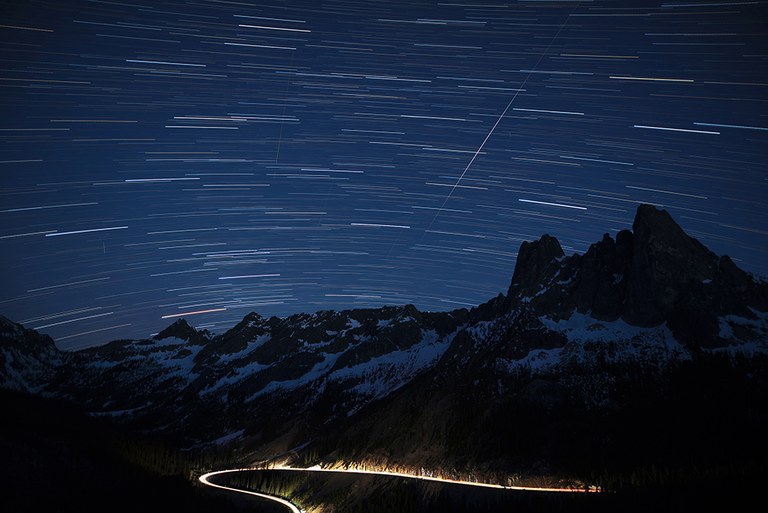 North Cascades Highway at night. Photo by Dan Clements. A starry sky and mountains with an illuminated highway curving around the base of the mountains.