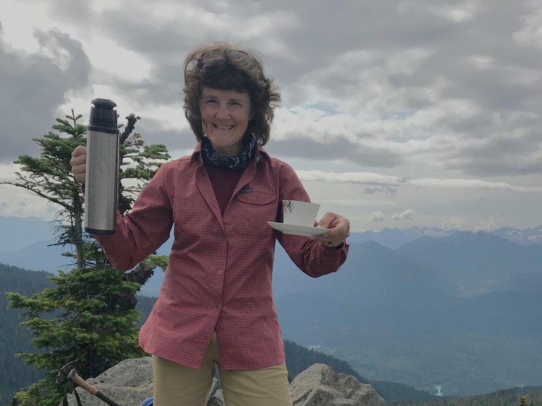 A hiker with short brown hair whipping in the wind holds a thermos and teacup