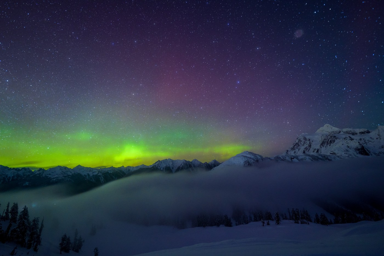 The northern lights glow on the horizon. Above twinkles a blanket of stars. Below sits dramatic mountaintops shrouded in mist.