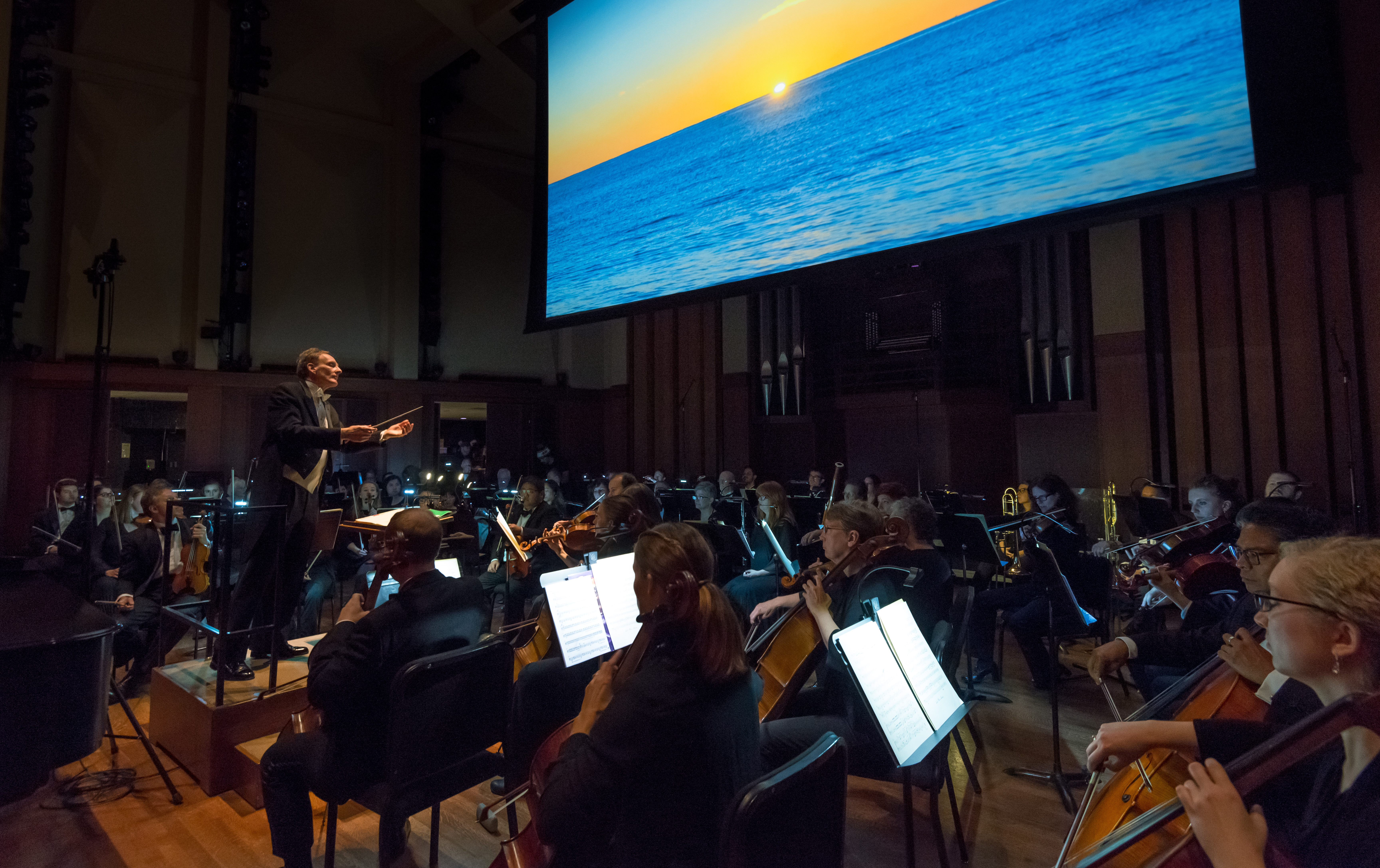 Symphony orchestra preforms in front of screen with photo of sunset on water.