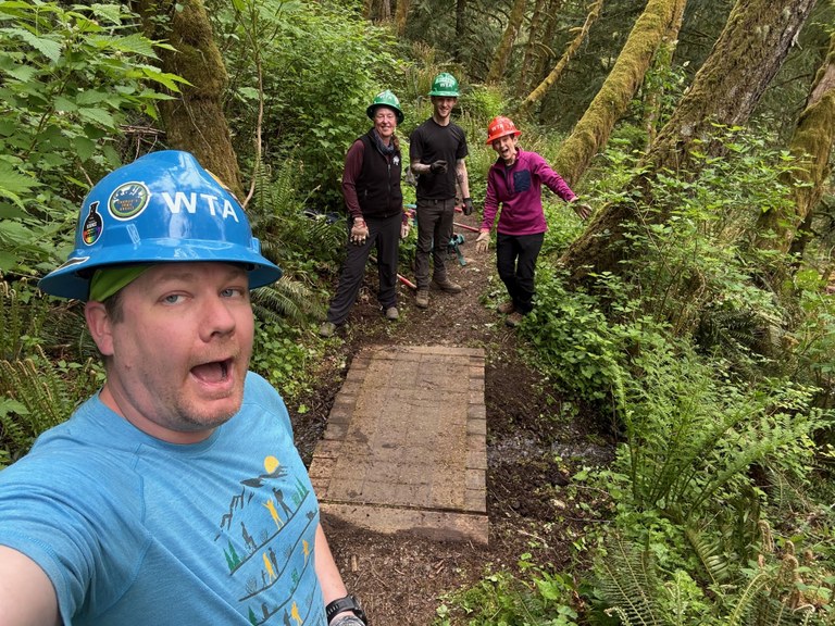 Man taking selfie with blue hard hat. Bridge in the background with three other people wearing hard hats smiling at camera in the woods.