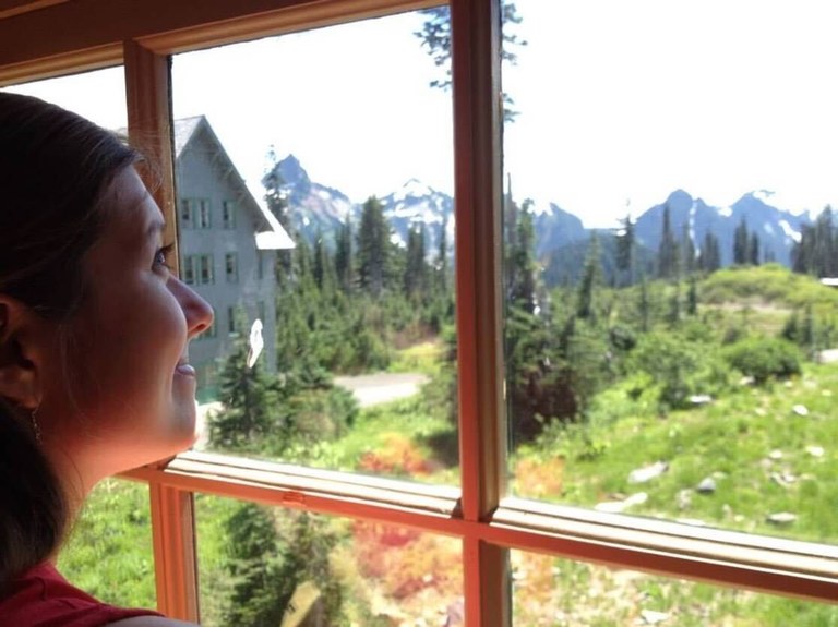 A yound white woman with brown hair smiles as she looks out the Paradise Lodge window at the Tatoosh mountain range. 