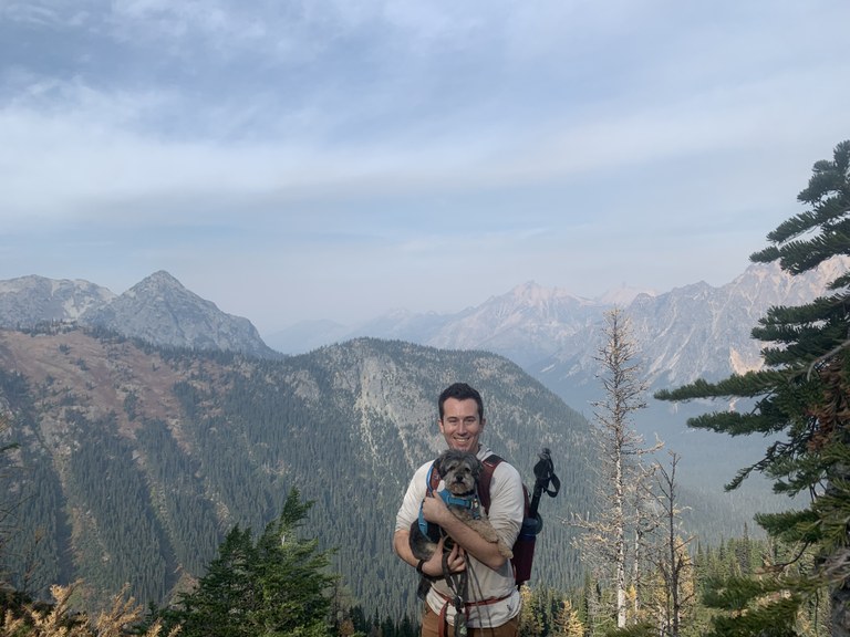 Hiker holding small dog in arms, smiling with mountains in the background. 