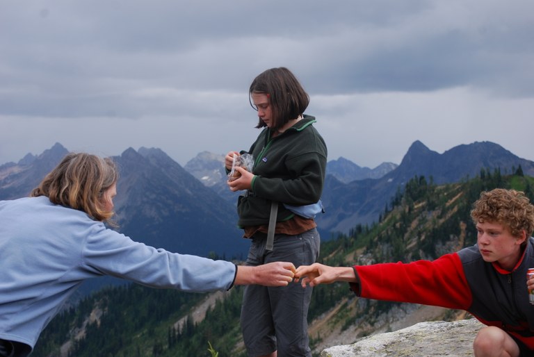 Two hikers swap snacks with outstreched arms while resting on trail. Mountains, grey skies and fellow hiker eating snack in background.