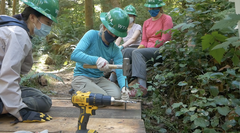 A teen volunteer uses a hammer while other volunteers watch. Everyone is wearing green hard hats and face coverings.