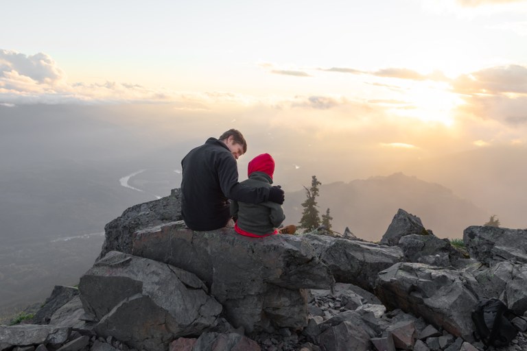 A man and child watch a beautiful sunset on a rock outcropping overlooking the mountains.