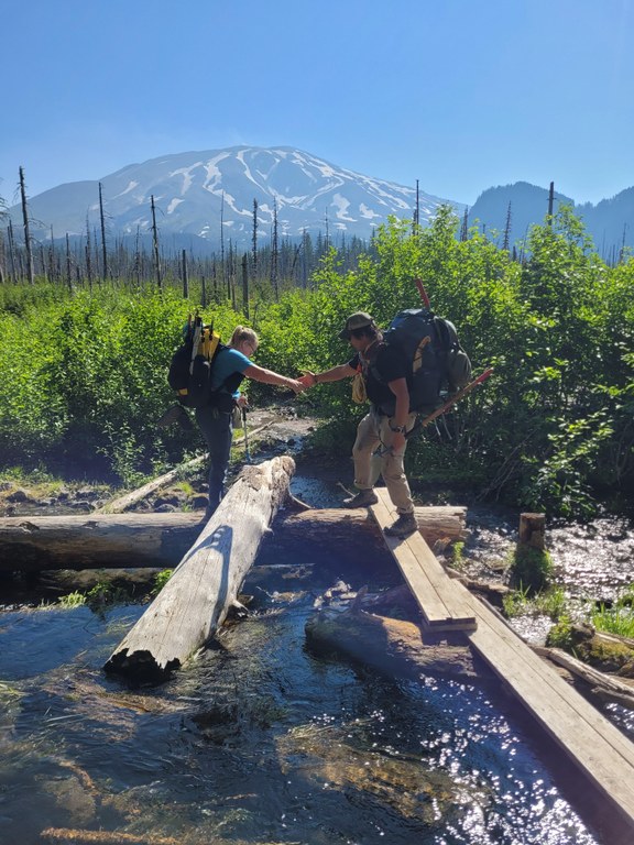 Two hikers wearing large packs with trail tools cross creek on log jam, on offering a helping hand from land with large snowy mountaina and burned trees in the background.