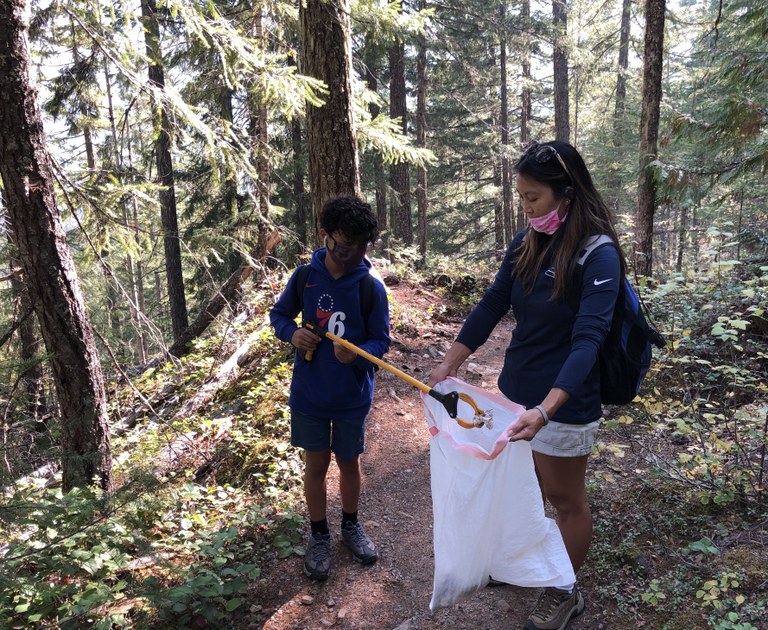 One person picks up trash on trail with long claw and other holds bag open for them. 