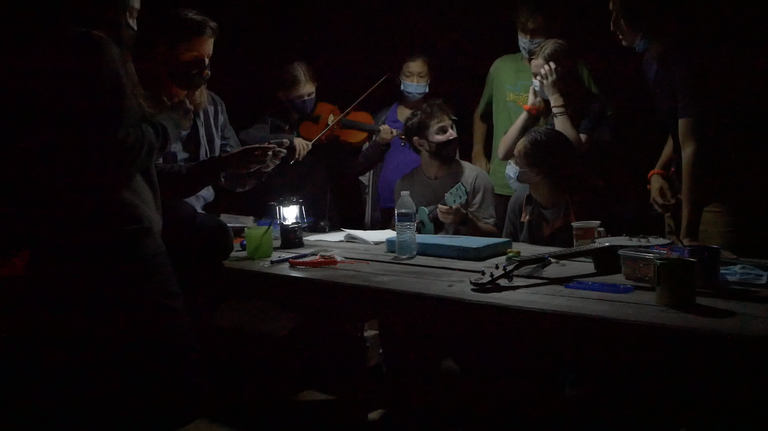 Teens stand around a picnic table in the dark, lit by a lantern. Teens are playing stringed instruments, and a few more instruments sit on the table. Everyone is wearing face coverings. 