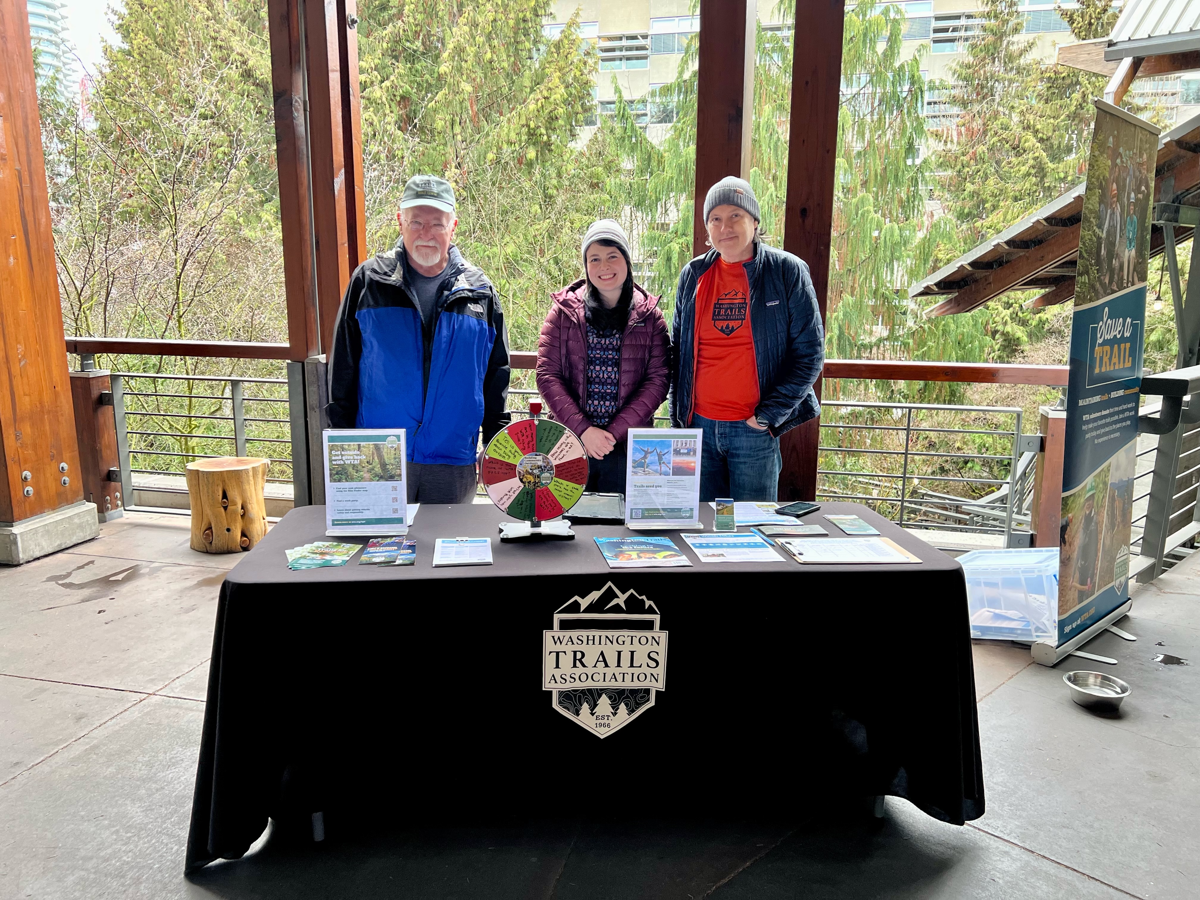 Three volunteers at tabling event at REI Seattle. Volunteers stand behind black table with WTA logo on the front. Trees in background of REI Seattle store.