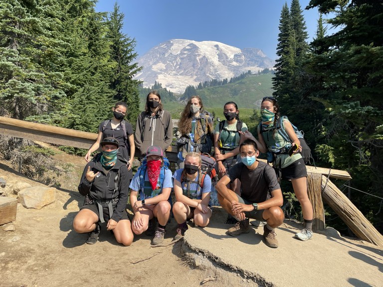 A group of nine teens, all wearing face coverings, pose for a photo in front of a wooden fence with Mount Rainier in the background on a sunny day. 