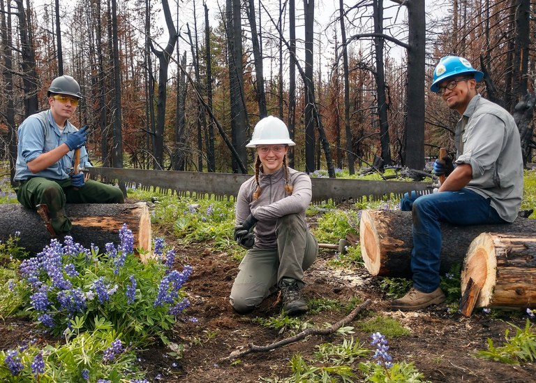 Three trail crew members pose for a photo on trail. The one in the center is wearing a white hat, the other two have green hats.