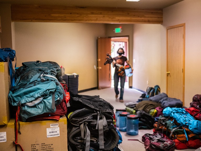 Puyallup gear library - photo by Lily Poppen A woman carrying a load of trekking poles and a bear canister walks into a room full of outdoor gear and boxes.