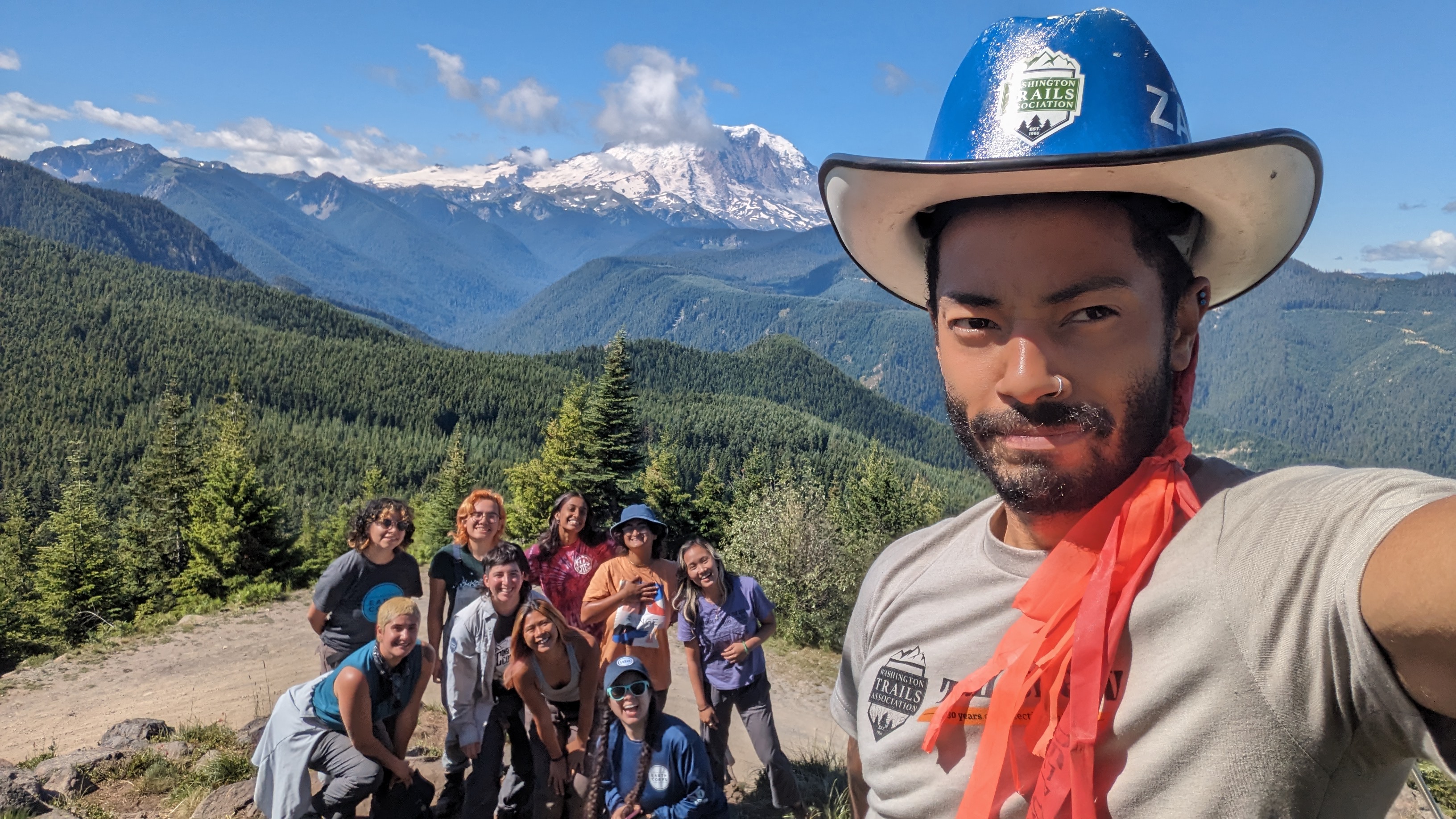 Zach Toliver, crew leader for WTA takes a selfie with a larger crew of BIPOC folks on the summit of a mountain 