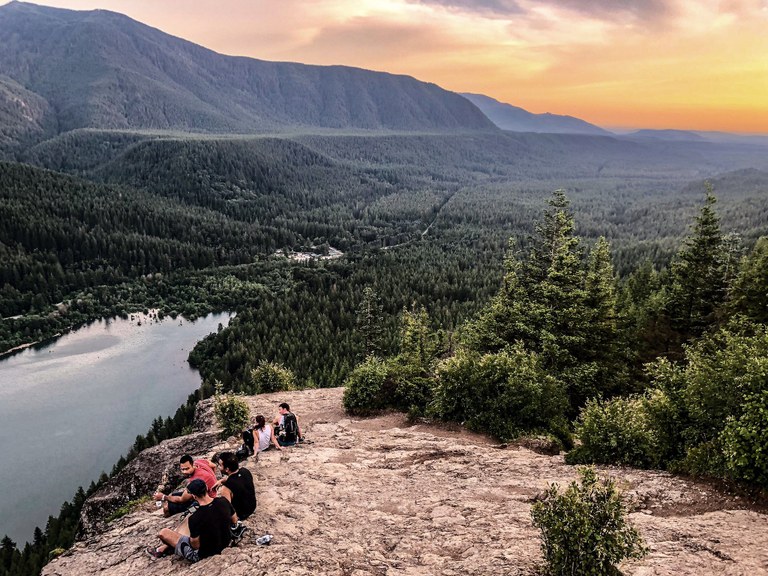 Several people sit on a rocky overlook above a lake with forest in the background at an orange sunset. Photo by Sean Downes. 