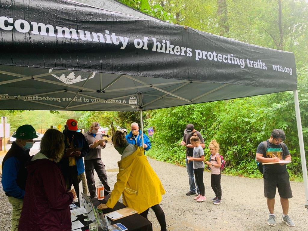Hikers in rain jackets sign postcards below a wet Washington Trails Association tent.