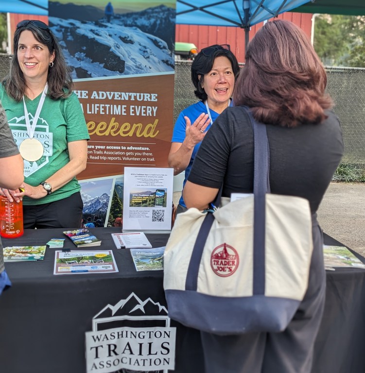 WTA representatives stand behind an outreach table at an outdoor event, engaged in conversation with attendees.