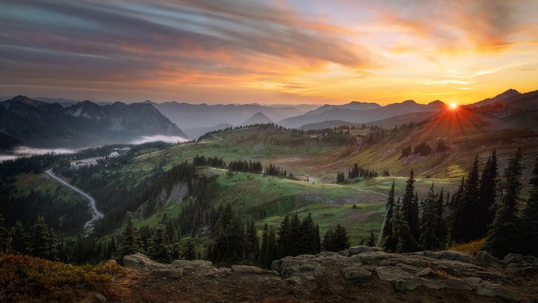 Sun setting over mountain ridges and green hills, with buildings and a winding road visible below. 