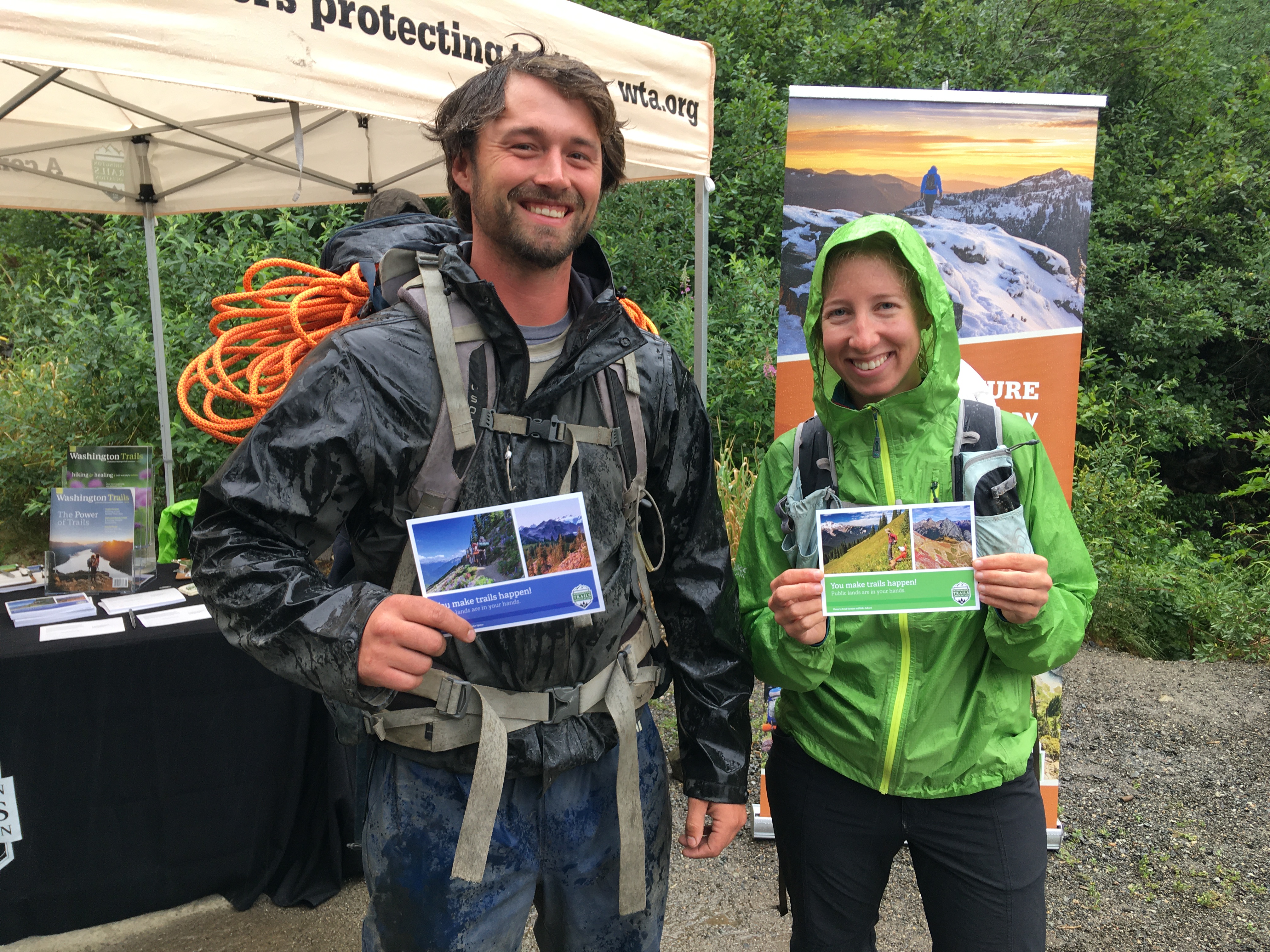 Hikers in rain jackets smile for the camera, holding their signed postcards for our Senators in front of them.