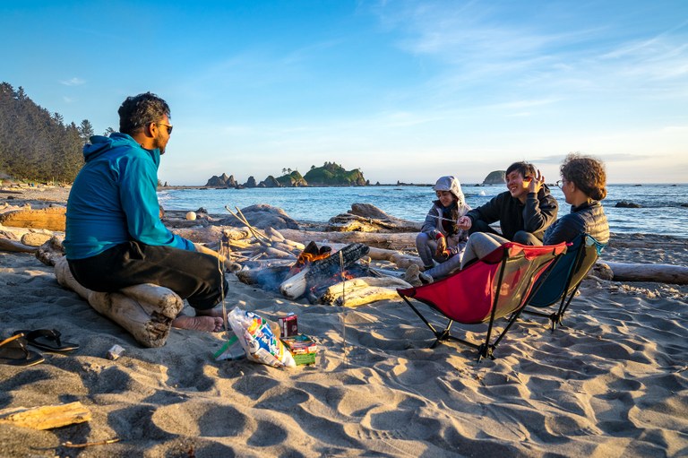 A group sits on chairs and driftwood around a small fire on the beach.