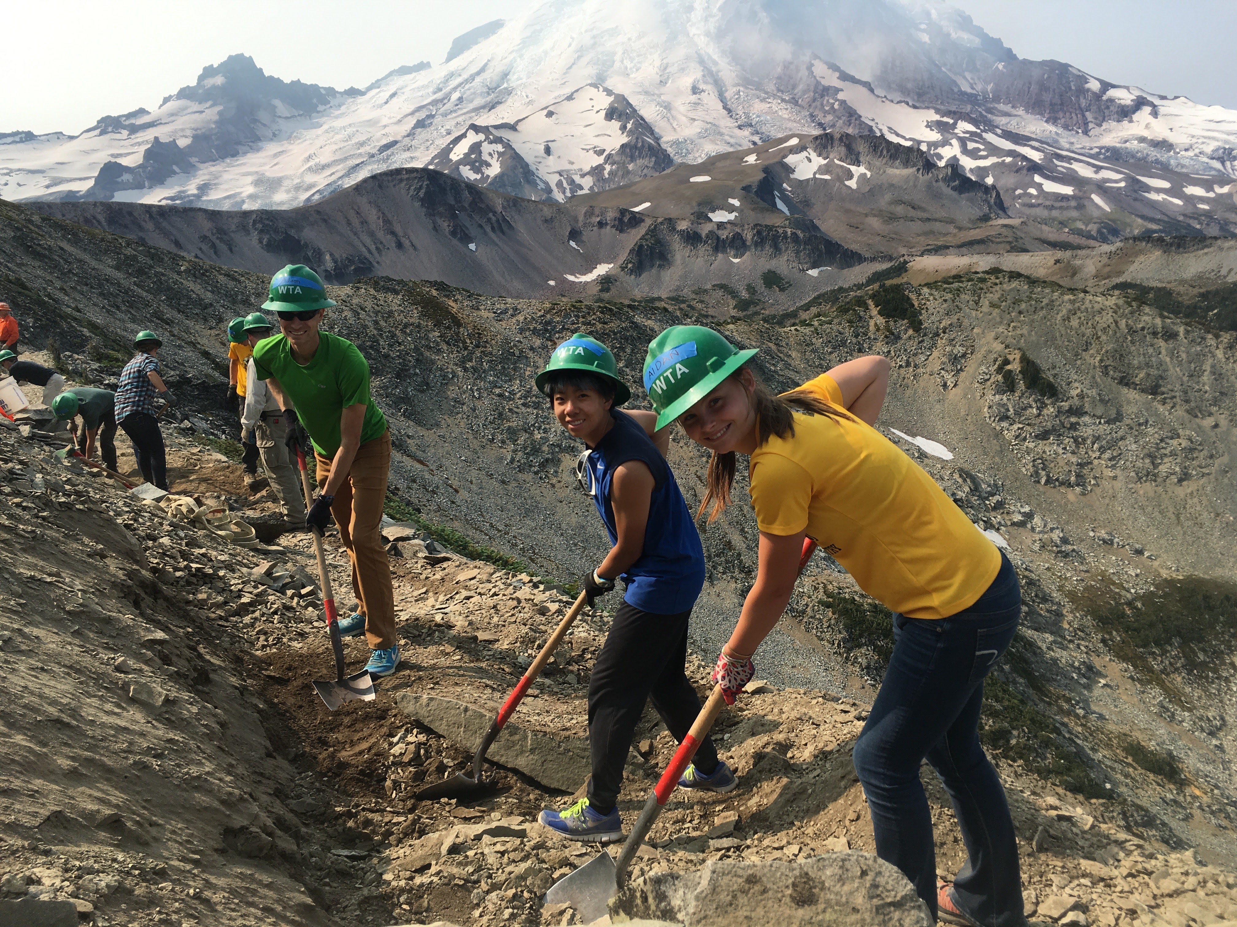 Trail crew members stop in the middle of digging a trail and smile for the camera. Mount Rainier creates an imposing background. 