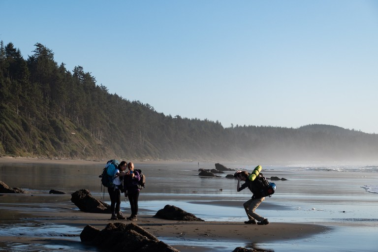 Man taking a photo of hikers. Photo by Mitchell Amundsen. Man with a backpack takes a photo of two hikers taking a selfie on the beach. Photo by Mitchell Amundsen.