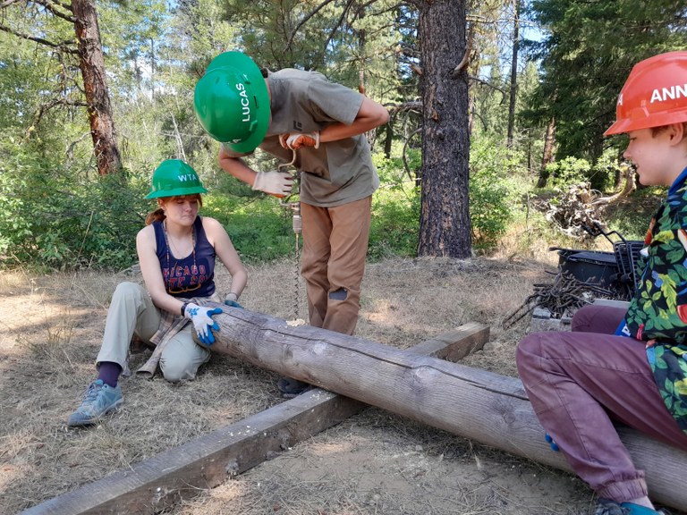 One youth volunteer uses a manual drill in a log while another volunteer and an assistant crew leader hold the log steady on a youth volunteer vacation in the Teanaway Community Forest. Photo by Sarah Williams.