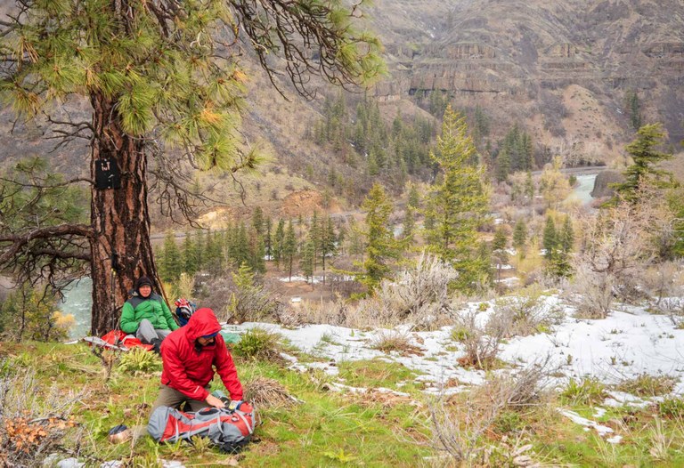 Two hikers take a break on the side of the trail. Patches of snow and pines surround them.