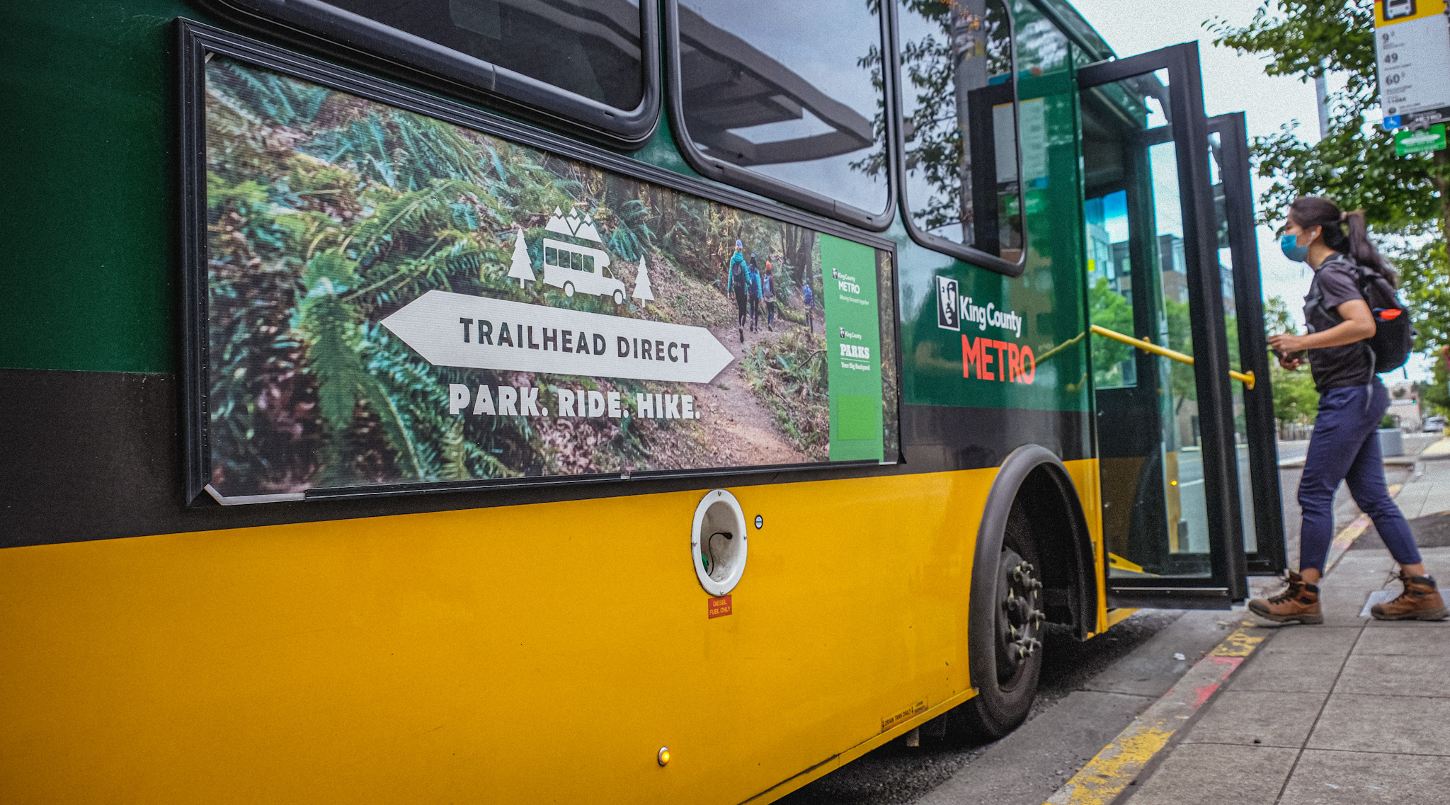 Person in boots boards bus with a sign that reads, "Trailhead Direct. Park. Ride. Hike."