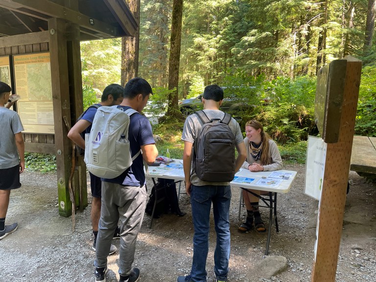 Three hikers look at a table of infromation about recreating responsibly while two trailhead ambassadors talk to them. 