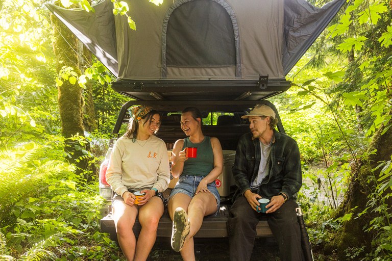 Three young hikers enjoy beverages in tin camp cups on the tailgate of a vehicle with a popup tent above. Photo by Carson Artec. 