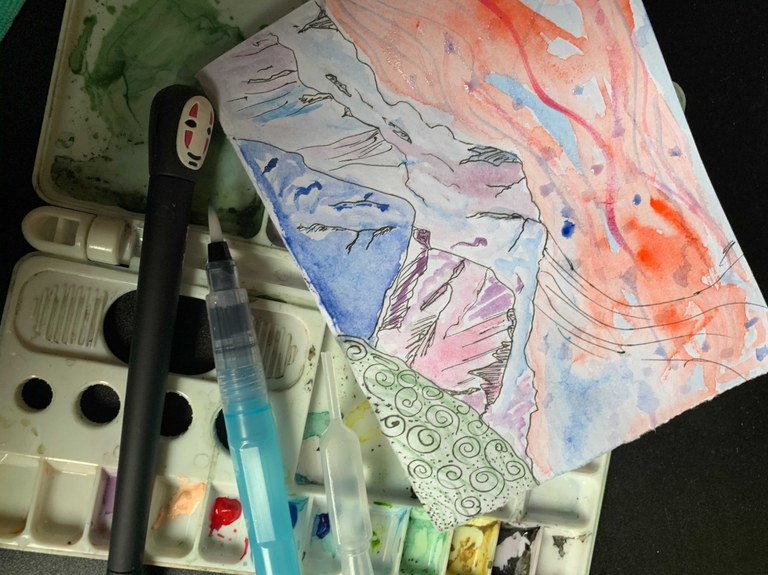 A set of watercolor paints, paintbrush and a painting of a mountain.