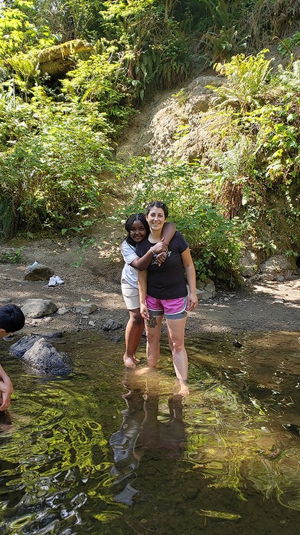 A woman stands in the stream with an older child behind her with her arms around her shoulders.