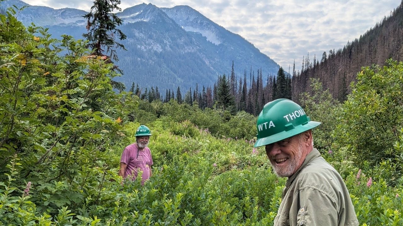 Trail crews at White River. Photo by Erin McMillan Two people in green hats smile at camera on trail covered in brush. Blue skies with clouds and mountains in the background.