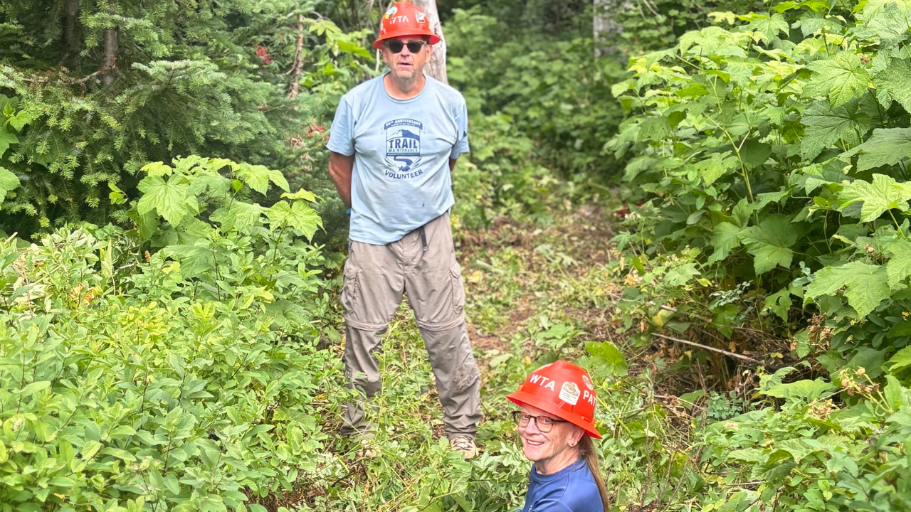 Two orange hats smile on trail. Photo by Erin McMillan. Two people wearing orange hats smile from trails covered in brush.