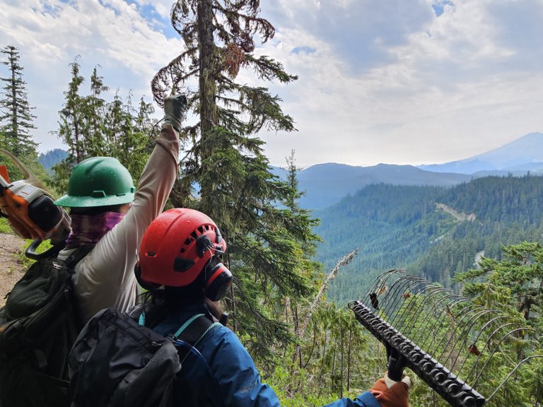 Volunteers happy on trail at St. Helens. Two people face away from the camera and overlook a lush green valley. One has his hand triumphanly in the air.