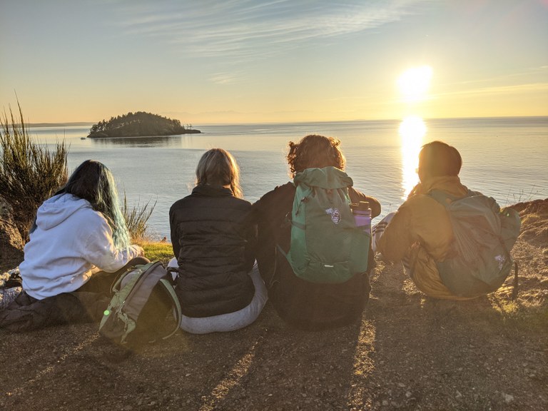 Four kids sit on a bluff overlooking saltwater as the sun sets. 